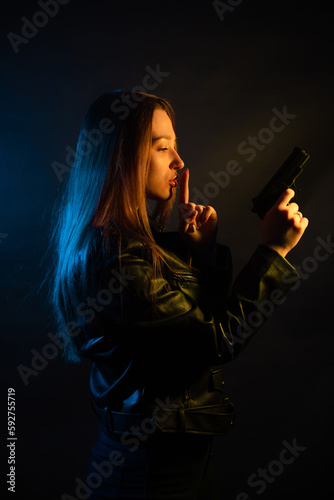 Beautiful young woman in the dark in a leather jacket with a gun in her hands. Girl in the club at night.