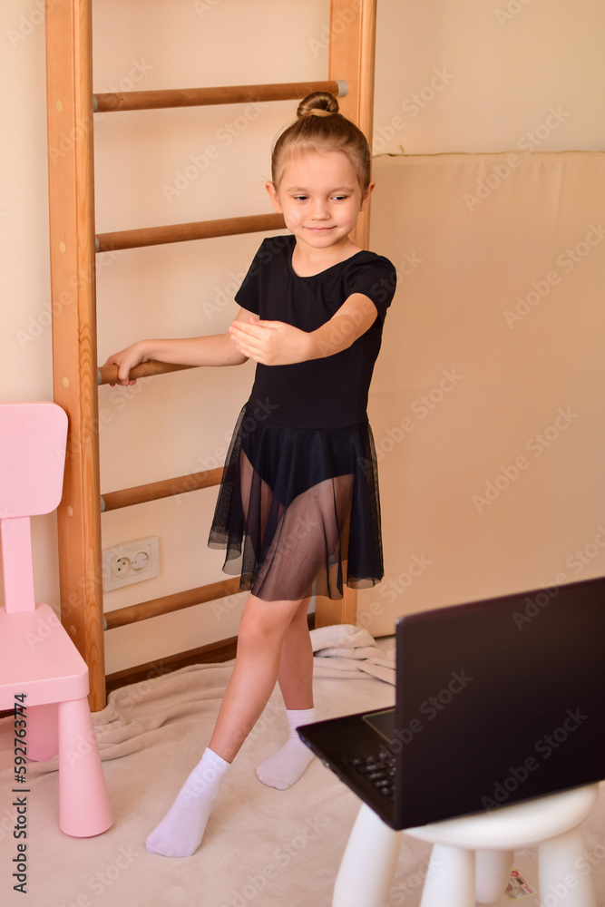 A little cute girl in a children's room in front of a laptop is engaged with a trainer via video communication with ballet, rhythmic gymnastics, yoga. The child undergoes online training at home. A little cute girl in a children's room in front of a laptop is engaged with a trainer via video communication with ballet, rhythmic gymnastics, yoga. The child undergoes online training at home.