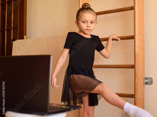 A little cute girl in a children's room in front of a laptop is engaged with a trainer via video communication with ballet, rhythmic gymnastics, yoga. The child undergoes online training at home.