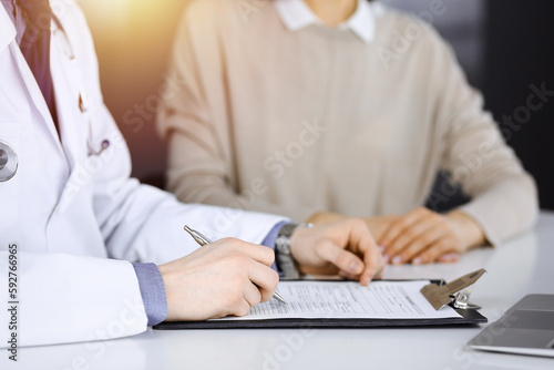 Unknown male doctor and patient woman discussing something while sittingin a darkened clinic and using clipboard  glare of light on the background