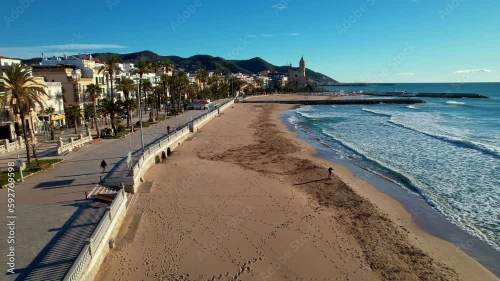 Panoramic drone aerial view of coastal city, old town of Sitges seen during sunny day. Close-up of surging Mediterranean sea waves. People are walking along the embankment. Spain. Film intro. 4K video