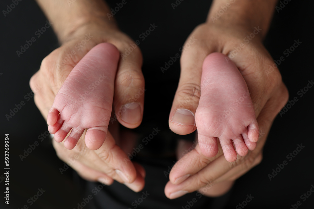 A mother hold the feet of a newborn child in a black blanket on a Black background. The feet of a newborn in the hands of parents. Studio macro photo legs, toes, feet and heels of a newborn. A mother hold the feet of a newborn child in a black blanket on a Black background. The feet of a newborn in the hands of parents. Studio macro photo legs, toes, feet and heels of a newborn.