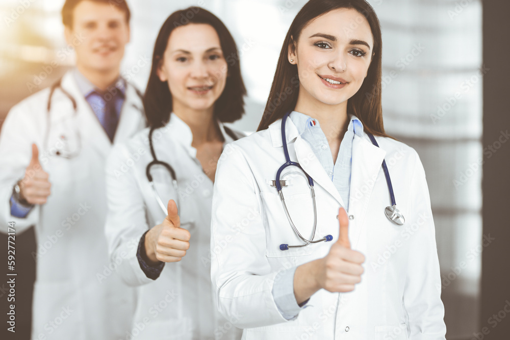 Group of young cheerful doctors standing as a team with thumbs up in sunny hospital. Medical help, insurance in health care, best disease treatment and medicine concept Group of young cheerful doctors standing as a team with thumbs up in sunny hospital. Medical help, insurance in health care, best disease treatment and medicine concept