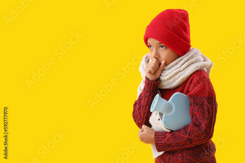 Fotografia Ill African-American boy with hot water bottle coughing on yellow background