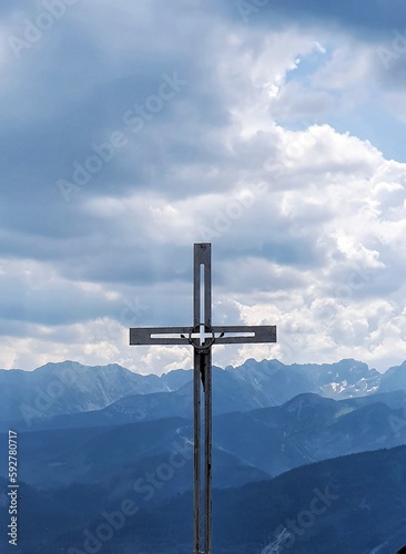 On the top of the hill, in the countryside of Poland, a metal cross facing the mountains on a day with blue sky and several clouds forming cotton balls.
