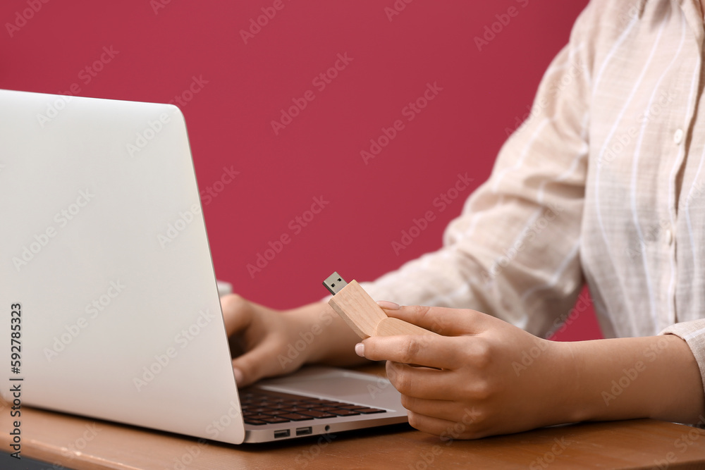 Woman using USB flash drive with modern laptop at wooden table near color wall, closeup