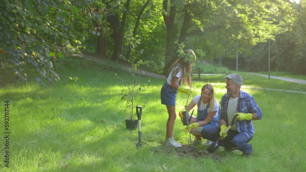 Cute family plant tree in the park together. Caucasian eco activists couple and their female child with shovels planting plant in soil. People and ecology concept. Cute family plant tree in the park together. Caucasian eco activists couple and their female child with shovels planting plant in soil. People and ecology concept.