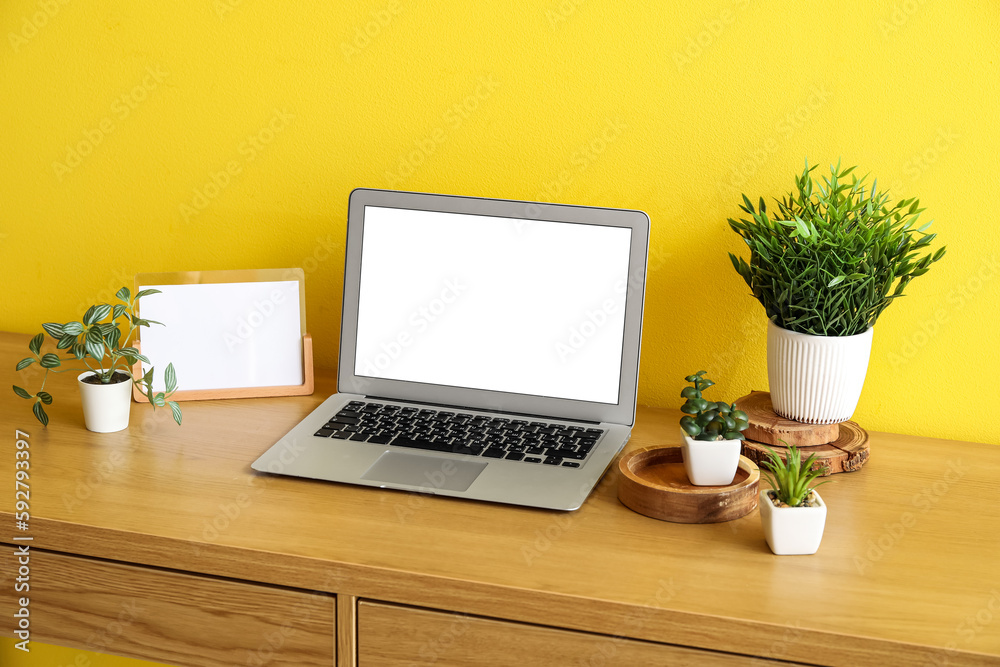 Workplace with artificial plants and laptop near yellow wall Workplace with artificial plants and laptop near yellow wall