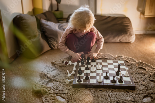 child plays single game of chess at home on floor. Chessboard with the end of the game of chess. Girl's hand moving a chess piece is partially visible.