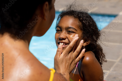 Biracial mother using suncream on happy daughter's face by swimming pool