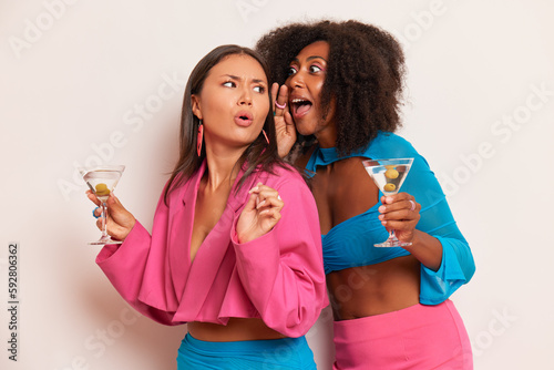 Two pretty ladies having party in studio, black girl in blue top whispers something to Asian girl in pink stylish jacket, both girls hold glass of cocktail, happy time concept, copy space, high