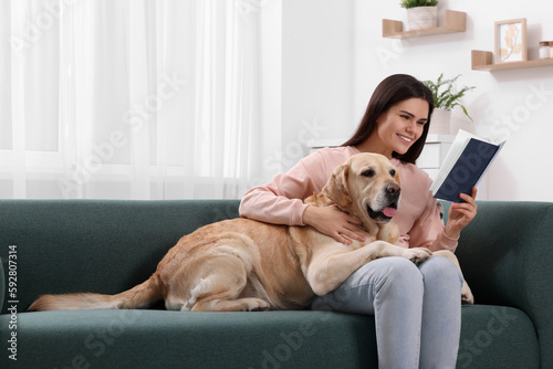 Happy woman reading book while sitting with cute Labrador Retriever on sofa at home