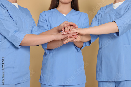 Nurses in medical uniforms putting hands together on light brown background, closeup