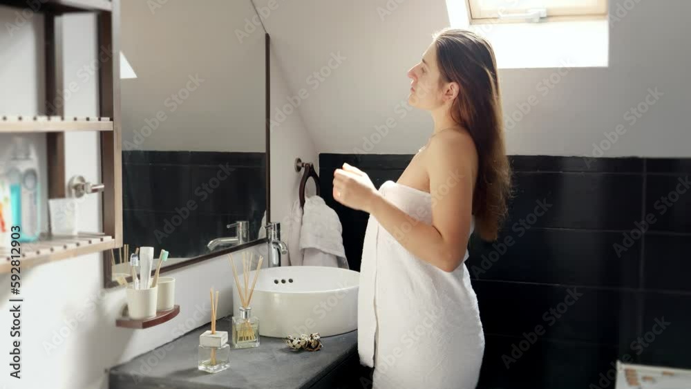 Woman covered in a towel, standing in a bath, and taking care of her long hair in front of a mirror. Female hygiene and self-care routines Woman covered in a towel, standing in a bath, and taking care of her long hair in front of a mirror. Female hygiene and self-care routines
