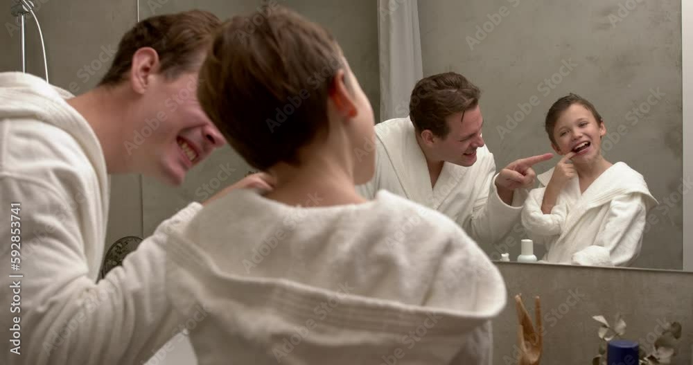 parent man with child boy in white bathrobes having morning routine in bathroom, standing near the mirror. Dad and kid checking tooth condition. Family vacation, healthcare concept. parent man with child boy in white bathrobes having morning routine in bathroom, standing near the mirror. Dad and kid checking tooth condition. Family vacation, healthcare concept.