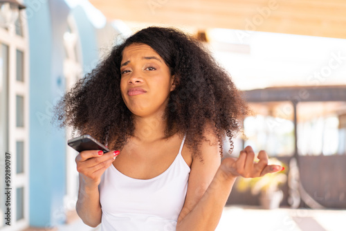 Young African American woman using mobile phone at outdoors making doubts gesture while lifting the shoulders