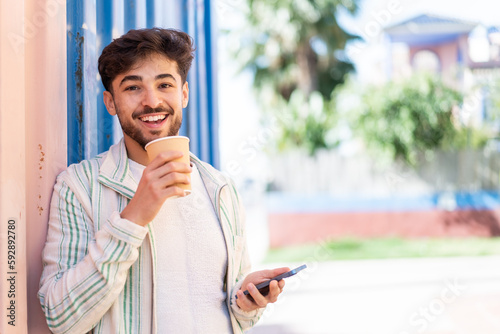 Handsome Arab man at outdoors using mobile phone and holding a coffee with happy expression