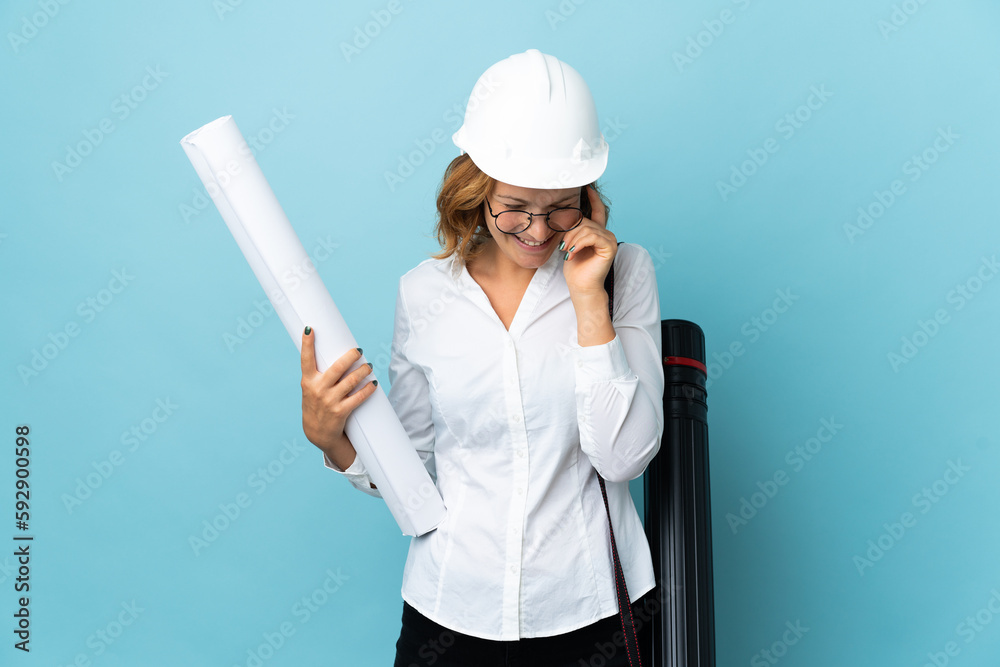 Young architect Georgian woman with helmet and holding blueprints over isolated background laughing Young architect Georgian woman with helmet and holding blueprints over isolated background laughing