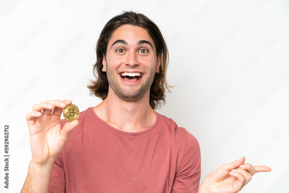 Young handsome man holding a Bitcoin isolated on white background surprised and pointing finger to the side Young handsome man holding a Bitcoin isolated on white background surprised and pointing finger to the side