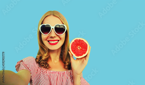 Summer portrait of happy smiling young woman posing stretching her hand for taking selfie with smartphone with juicy slice of grapefruit fruit wearing heart shaped sunglasses on blue background