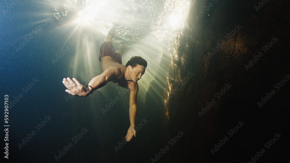 Man dives in the freshwater river with sunny rays shining through the water and swims near the rock