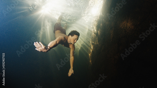 Man dives in the freshwater river with sunny rays shining through the water and swims near the rock