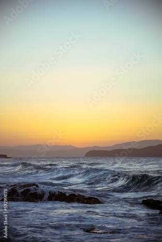Vertical shot of sea waves on the shore with mountain silhouettes on the horizon during sunset © Frajobs/Wirestock Creators