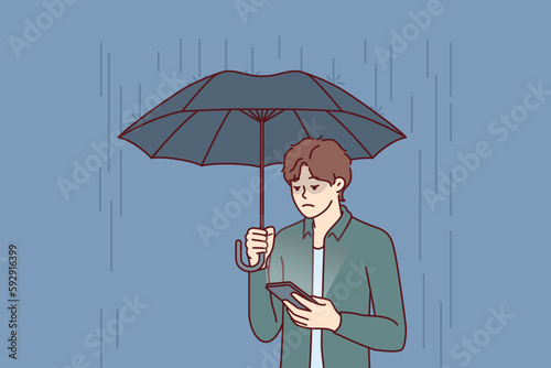 Sad man with umbrella stands in rain and reads SMS in mobile phone from girlfriend who refused to come on date. Concept of autumn depression due to bad weather and weather sensitivity