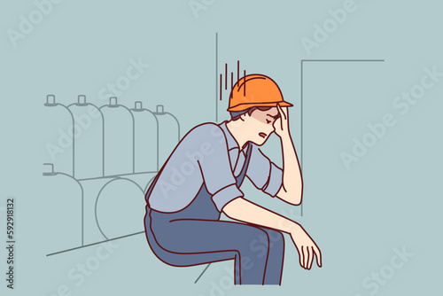 Tired man working in factory sits near production equipment holding head because of nervous job. Nervous factory worker in safety helmet needs psychological support due to poor working conditions