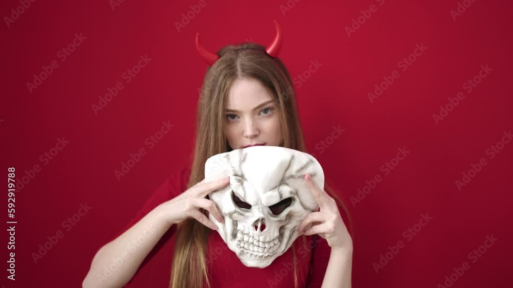 Young blonde woman wearing devil costume holding skull mask over isolated red background Young blonde woman wearing devil costume holding skull mask over isolated red background