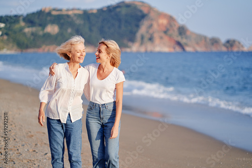 Outdoor portrait of smiling happy caucasian senior mother with her adult daughter hugging and walking on sea beach.
