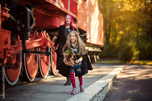 A woman accompanies her child to the school of magic. A girl in a raincoat, in a scarf, with a suitcase near the red train. Photo with a locomotive.