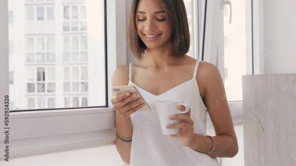 Woman dressed in white pajamas. Carefree female holds cup and drinking coffee. Sexy model enjoying her morning breakfast at balcony.Looking at cellphone screen using mobile apps Woman dressed in white pajamas. Carefree female holds cup and drinking coffee. Sexy model enjoying her morning breakfast at balcony.Looking at cellphone screen using mobile apps