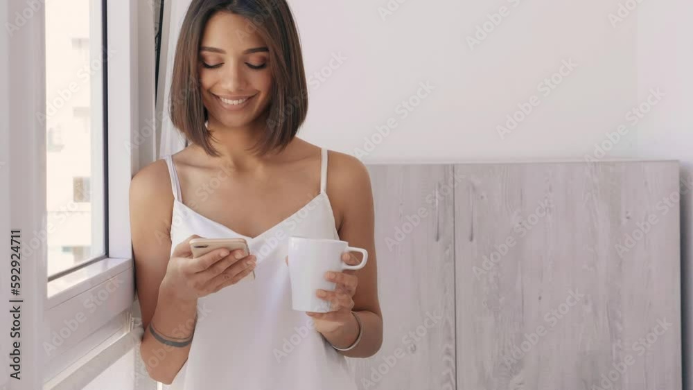 Woman dressed in white pajamas. Carefree female holds cup and drinking coffee. Sexy model enjoying her morning breakfast at balcony.Looking at cellphone screen using mobile apps Woman dressed in white pajamas. Carefree female holds cup and drinking coffee. Sexy model enjoying her morning breakfast at balcony.Looking at cellphone screen using mobile apps