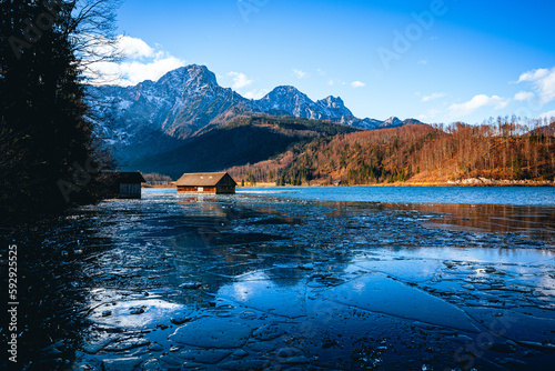 Distant shot of a wooden house in the middle of a frozen lake, colorful trees and rocky mountains