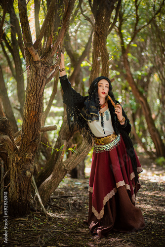 A gypsy woman in the forest with a wooden smoking pipe in her hands. The black-haired witch conjures in the forest. Women s Magic.