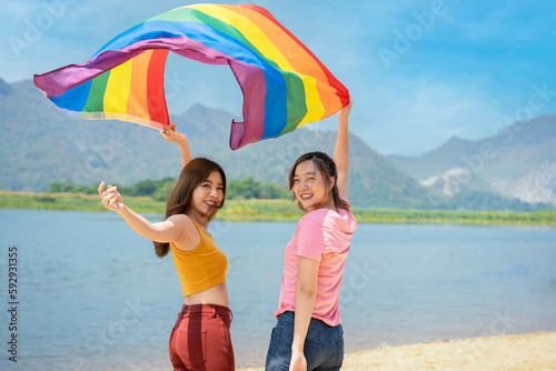 Happy lesbian, beautiful asian young two women, girl gay, couple love moment spending good time together, holding or waving lgbt rainbow, pride flag  together on the beach.