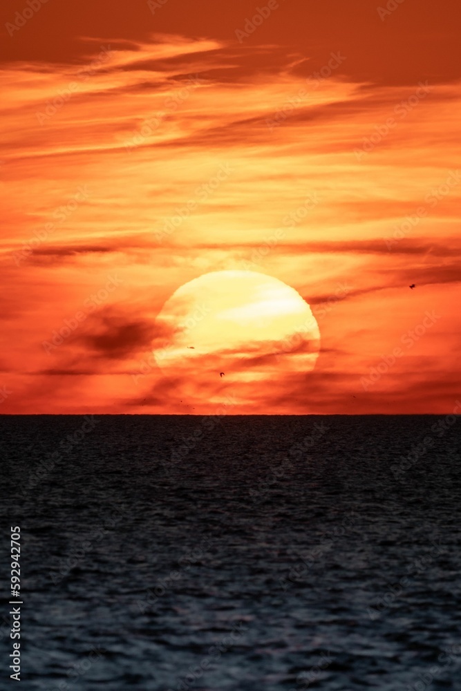 Vertical of a big glowing sun captured in the red sunset sky above a tranquil evening seascape