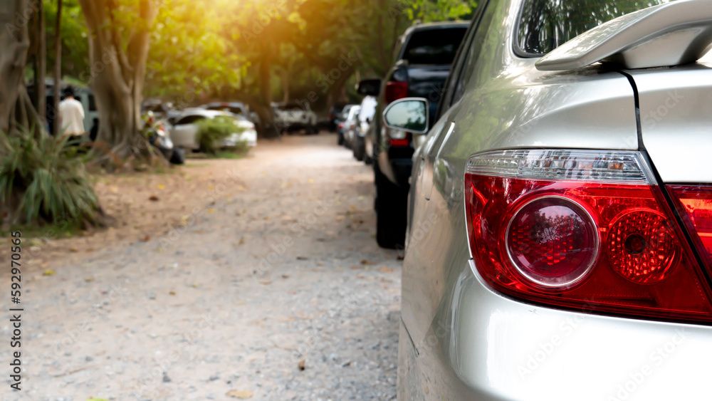 Rear view of a car in a residential parking lot. Visible taillights of car and blurred of mirror wing. Long row of cars parked in quiet neighborhood. Blurred of shade of tall trees beside road.