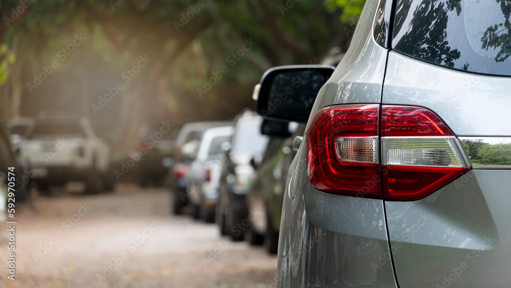 Rear view of a car in a residential parking lot. Visible taillights of a gray car and blurred of mirror wing. Long row of cars parked in quiet neighborhood. Blurred of shade of tall trees beside road.