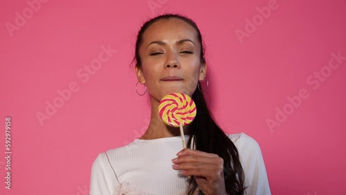 Closeup portrait of sexy girl eating lollipop. Woman tongue licking candy. Model dressed in summer hipster clothes. Isolated over pink background photo