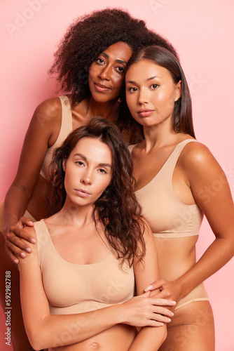 Photo portrait of different skin girlfriends dressed in nude-colored underwear, one girl sits in front, her friends stand behind hugging, friendship concept, copy space, high quality photo © South House Studio