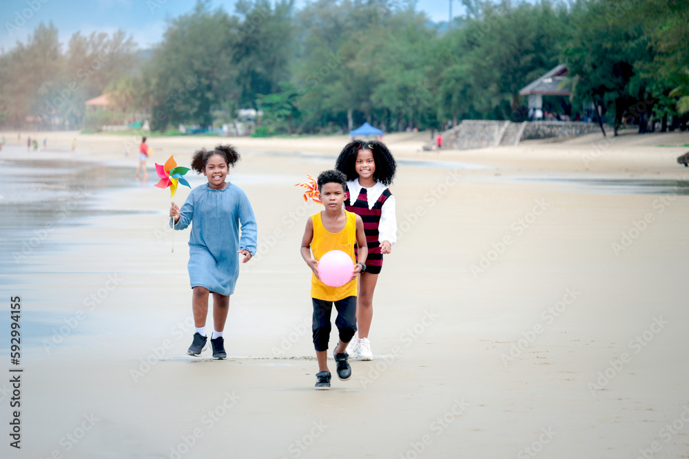 Obraz premium Cute groups of African kids have fun activity together on summer beach blue sea, two girls with black curly hair and boy running on tropical beach, happy childhood friends on holiday vacation.