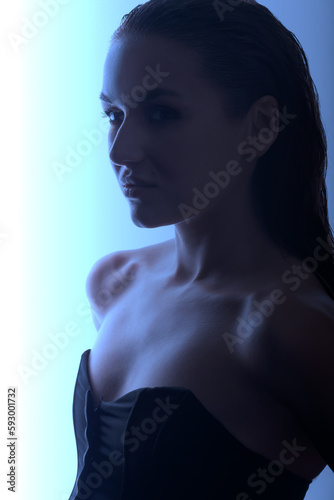 Portrait of beautiful and sexy brunette woman silhouette with black corset and wet hair. Model standing in grace pose and looking aside camera with seductive look. Toned image with blue color