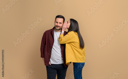 Young girlfriend whispering her desires into handsome boyfriend's ear while standing isolated over beige background. Attractive couple dressed in casuals gossiping together