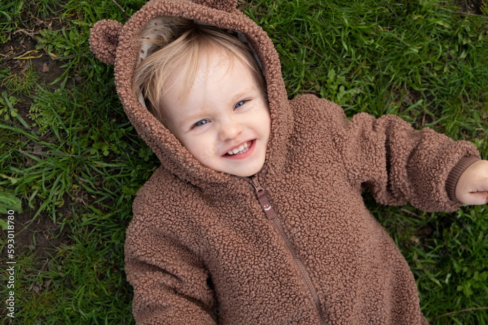 Portrait of a one-and-a-half-year-old blue-eyed girl in a bear costume lying on the grass, view from above Portrait of a one-and-a-half-year-old blue-eyed girl in a bear costume lying on the grass, view from above