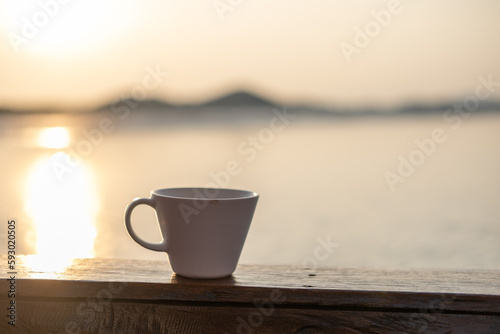 A cup of coffee on the wooden deck, with sunrise and sea view in the background