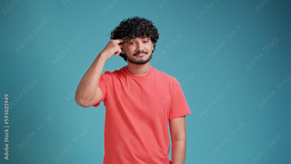 Indian man pointing his finger at his head showing high intellect and good brains and ability to think in coral t-shirt on blue studio background. Indian man pointing his finger at his head showing high intellect and good brains and ability to think in coral t-shirt on blue studio background.