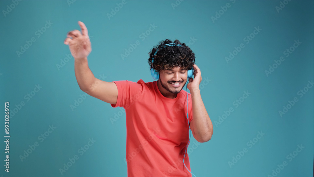 Young smiling happy cheerful student Indian man 20s dancing with headphones listen to music have fun in coral t-shirt on blue studio background. People lifestyle concept Young smiling happy cheerful student Indian man 20s dancing with headphones listen to music have fun in coral t-shirt on blue studio background. People lifestyle concept