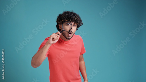 Funny Indian man looking through magnifying glass, searching or investigating something, standing in coral t-shirt on blue studio background